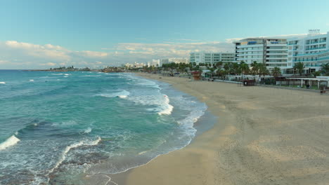 Vista-Aérea-De-La-Costa-De-Protaras,-Chipre,-Con-Playas-De-Arena,-Aguas-Cristalinas-Y-Un-Animado-Paseo-Marítimo-Bajo-Un-Cielo-Parcialmente-Nublado.