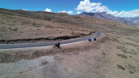 Hombre-De-Pie-Junto-A-La-Motocicleta-En-Un-Desierto-Paisaje-Montañoso-Seco,-Antena