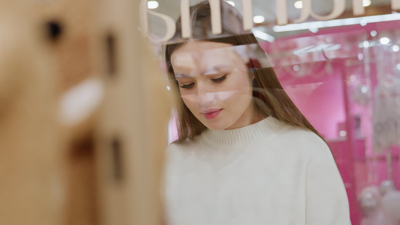 Close-up of a soft toy with a young woman admiring it through the glass, while people walk in the background in a bustling mall, creating a lively shopping atmosphere
