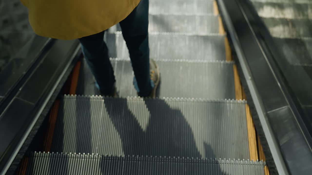 A close-up capturing the legs of a person wearing blue jeans and sneakers as they descend an escalator. The focus is on the movement and detail of the escalator steps in a busy mall setting