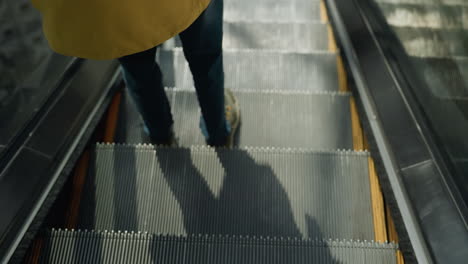 a close-up capturing the legs of a person wearing blue jeans and sneakers as they descend an escalator. the focus is on the movement and detail of the escalator steps in a busy mall setting