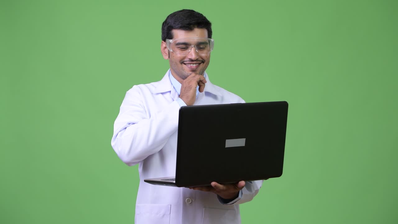 Young handsome Persian man doctor wearing protective glasses using laptop