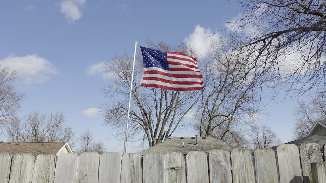 Una bandiera americana sventola dietro una staccionata di legno nel cortile di una casa ad Altoona, Iowa. I venti invernali fanno ondeggiare gli alberi spogli sotto un cielo grigio, creando una scena fredda e patriottica nel tranquillo Midwest.