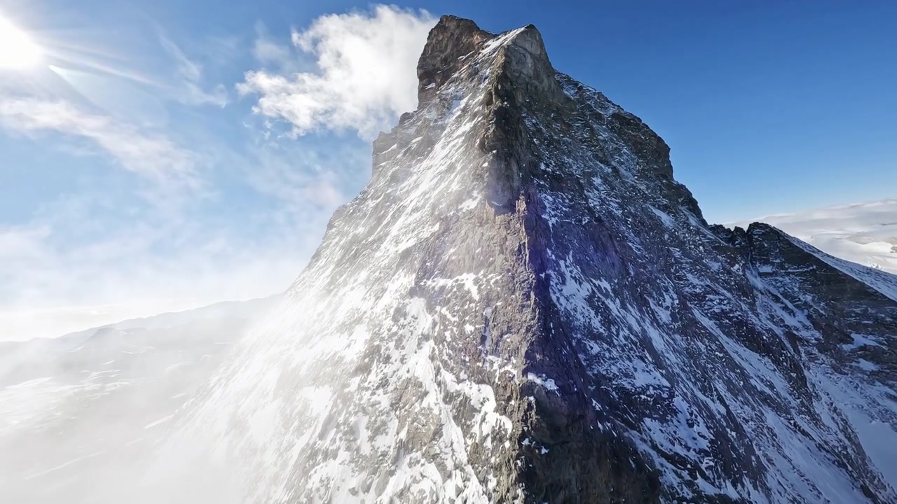 Volo drammatico con drone FPV che segue la cresta sommitale innevata del Cervino in Svizzera. Vedute aeree cinematografiche rivelano l'iconica vetta alpina, i ghiacciai e il terreno montuoso aspro.