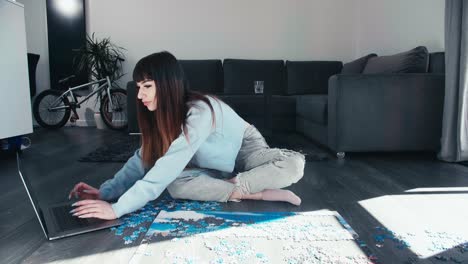 young woman sitting cross legged chatting on laptop while play with puzzle in modern flat house, girl sit cross legged in trendy clothes spend leisure time wisely, modern smart eco friendly apartment