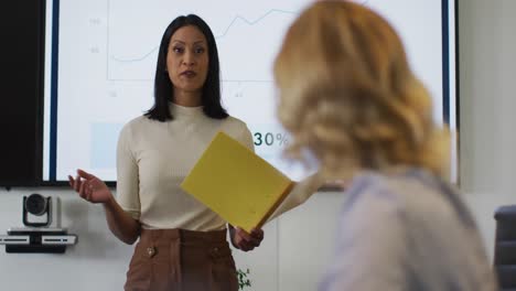 Mixed-race-businesswoman-giving-presentation-in-meeting-room-holding-documents-smiling