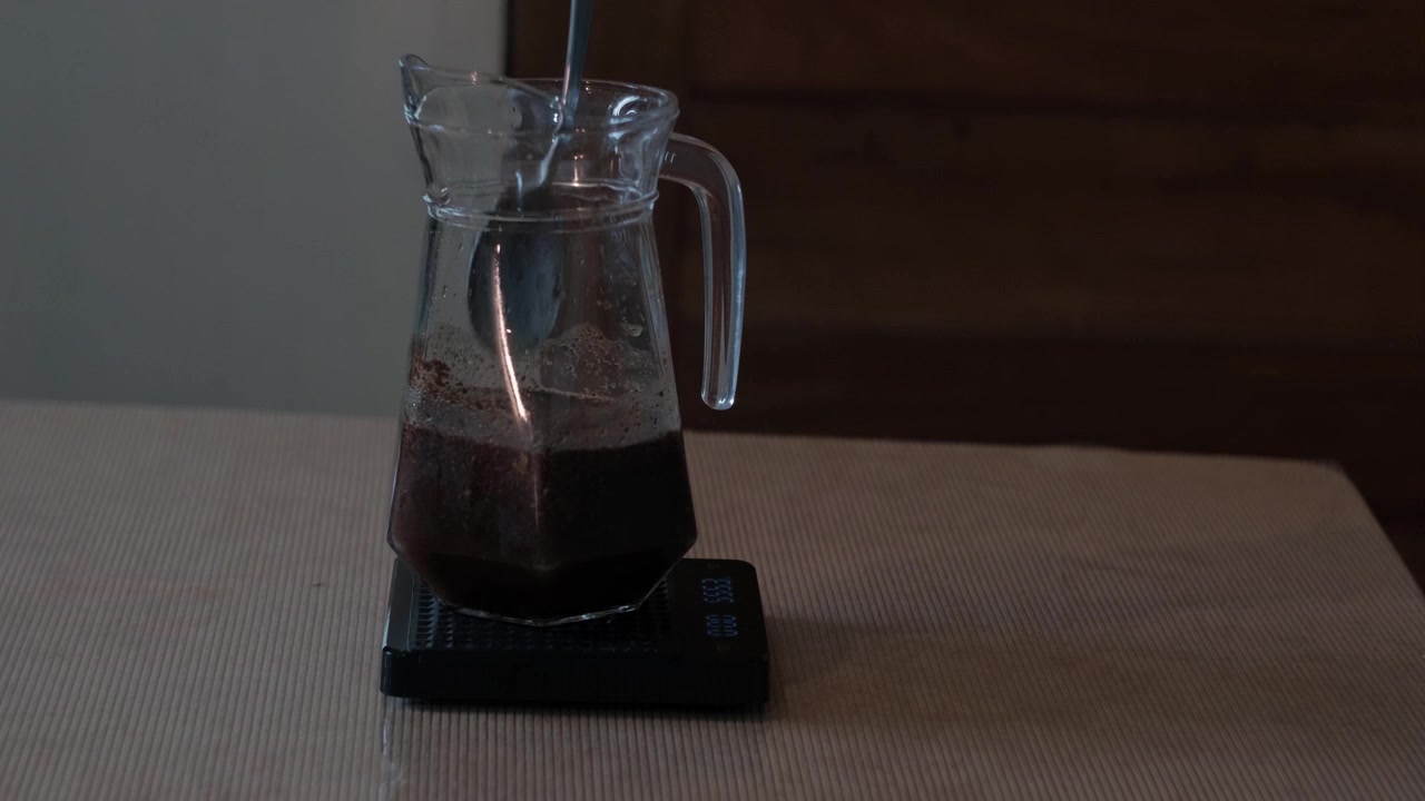 Inquadratura statica media di fondi di caffè e acqua fredda mescolati in una brocca di vetro, mentre si prepara una miscela per cold brew al chiuso prima dell'infusione