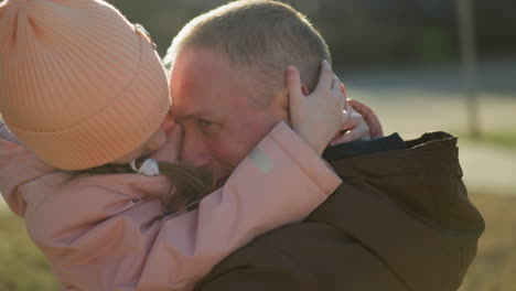 a touching moment captured as a little girl in a pink cap and jacket places her hands lovingly on a man's face while being carried. the sunlight adds a warm glow to this heartfelt interaction