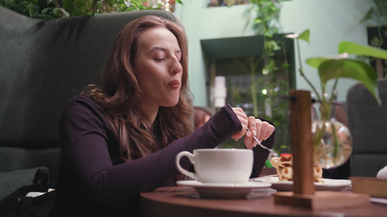 Lady enjoys waffles and sips latte in cozy cafeteria surrounded by greenery, with decorative vase in foreground and blurred seated guests in background adding depth to relaxed dining atmosphere