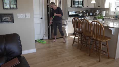 senior woman dusting and sweeping hardwood oak floors in the kitchen - tilt down