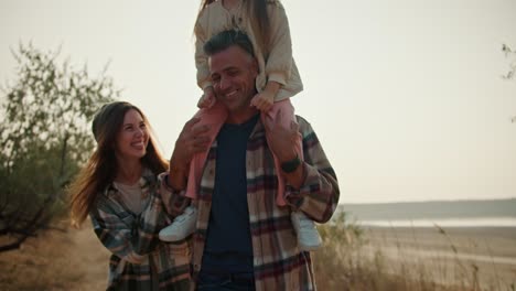 A-little-blonde-girl-is-having-fun-while-sitting-on-the-shoulders-of-her-brunette-dad-in-a-brown-checkered-shirt-who-is-running-with-his-wife-while-walking-along-a-path-outside-the-city