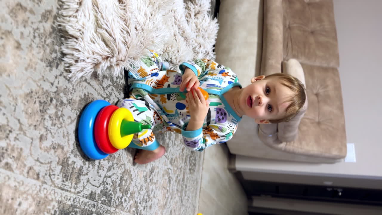 Beautiful Caucasian baby boy in colorful romper sits on the floor. Kid plays with a toy and holds his hand up for something. Vertical video.