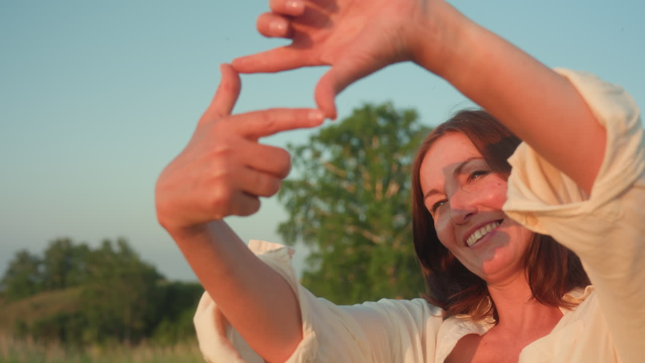 Close up of smiling woman framing sunlight with hand gesture, sun rays streaming through fingers, blurred river valley and hill backdrop illuminated by warm sunset glow
