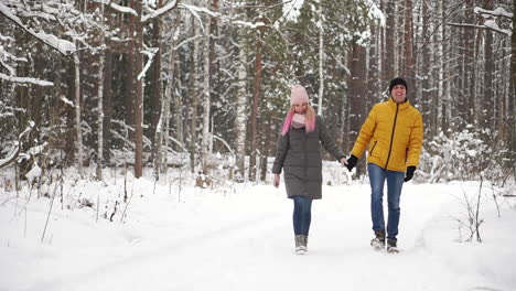 A-man-in-a-yellow-jacket-and-a-girl-in-a-hat-and-scarf-walk-through-the-winter-forest-during-a-snowfall-laughing-and-smiling-at-each-other-at-Christmas-in-slow-motion