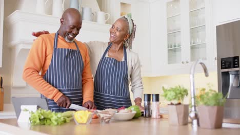 African-american-senior-couple-in-aprons-preparing-meal-using-tablet-in-kitchen,-slow-motion