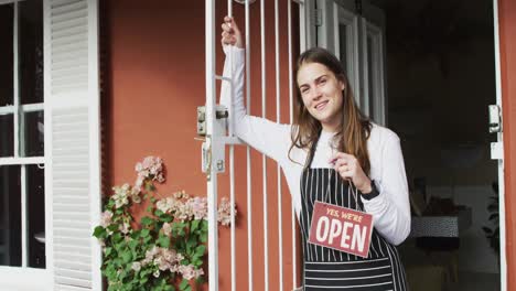 una camarera caucásica sonriente de pie en la puerta, sosteniendo un cartel abierto, mirando a la cámara