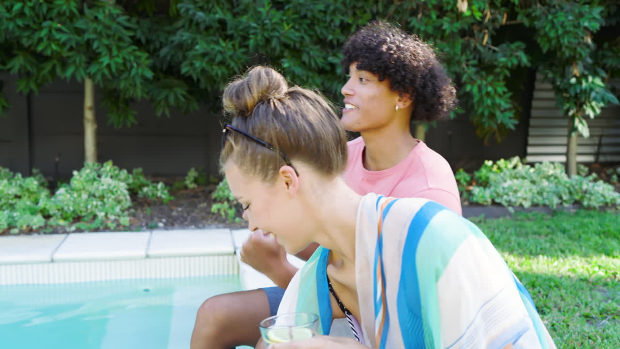 Diverse couple relaxing by pool, enjoying drinks and sharing joyful conversation outdoors