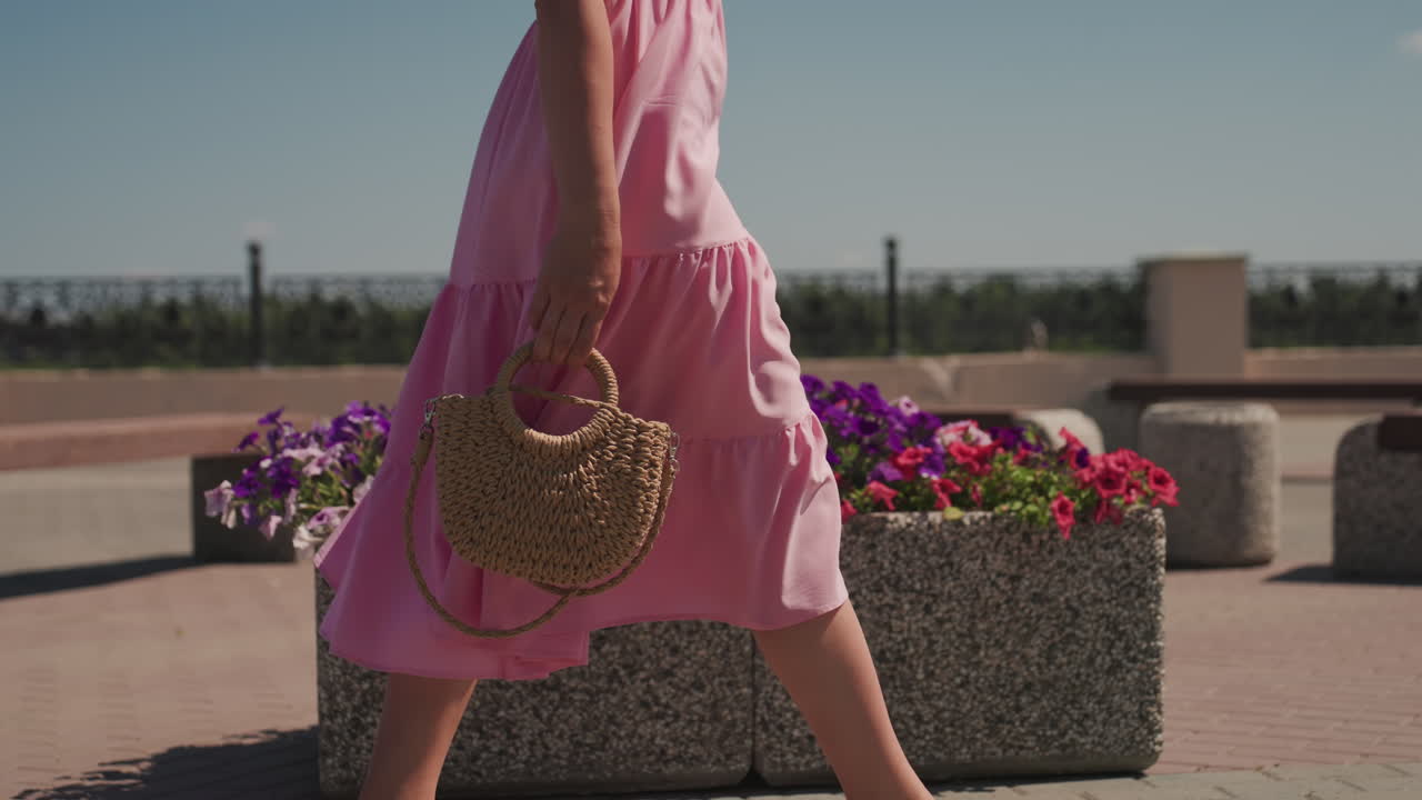 Donna con abito rosa che cammina sulla terrazza, passeggiata urbana illuminata dal sole con panchine in cemento e fioriere colorate, borsa di vimini che oscilla di lato, leggera brezza estiva che solleva l'orlo, moda casual elegante