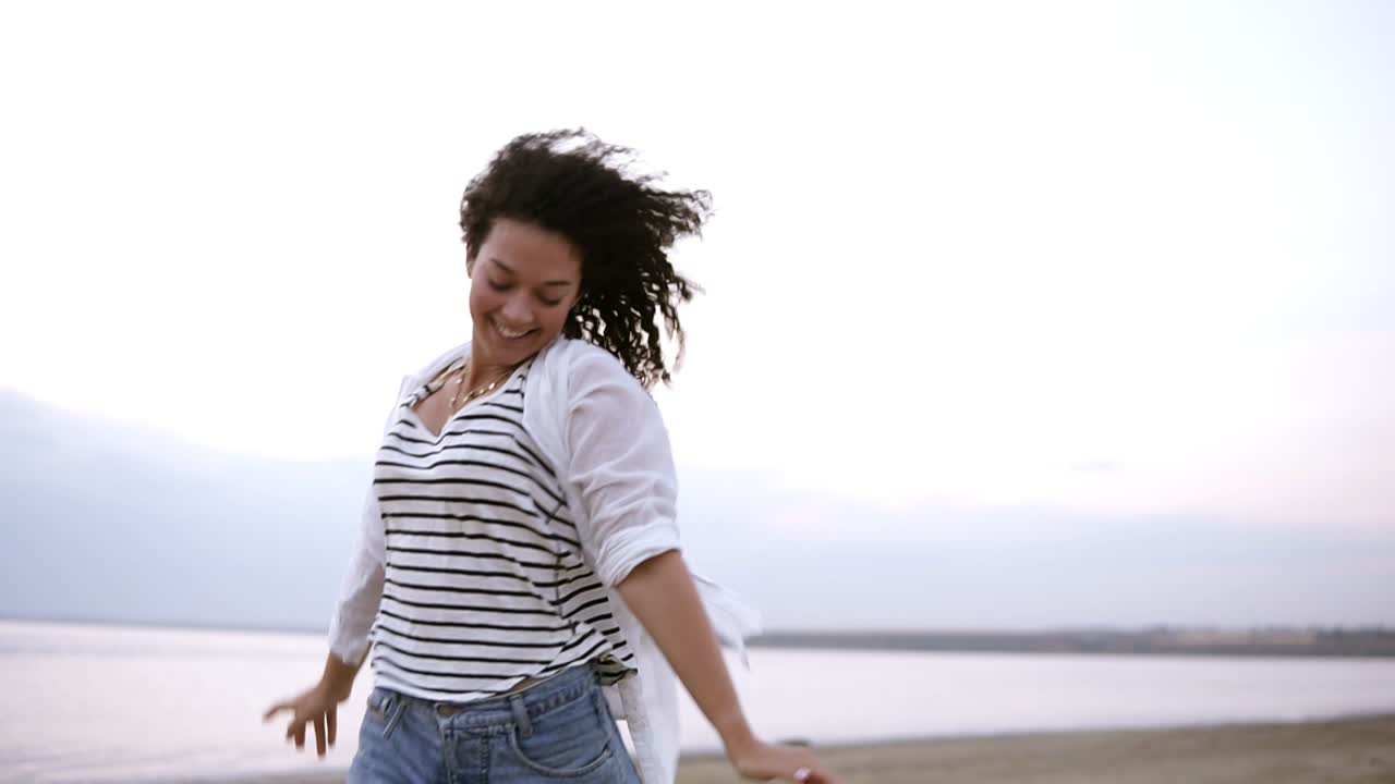 Portrait of a gorgeous curly brunette happily running near the sea or lake with outstretched hands. Wearing white casual clothes. Smiling, happy, freedom