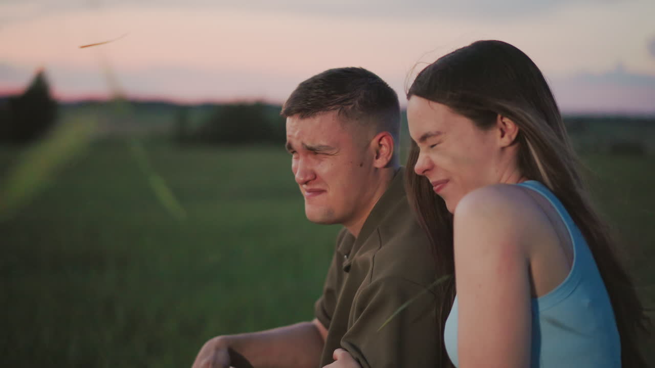 close up of woman holding man arm across eyes as cut grass flies around in windy field at sunset capturing playful protective gesture with serene landscape background
