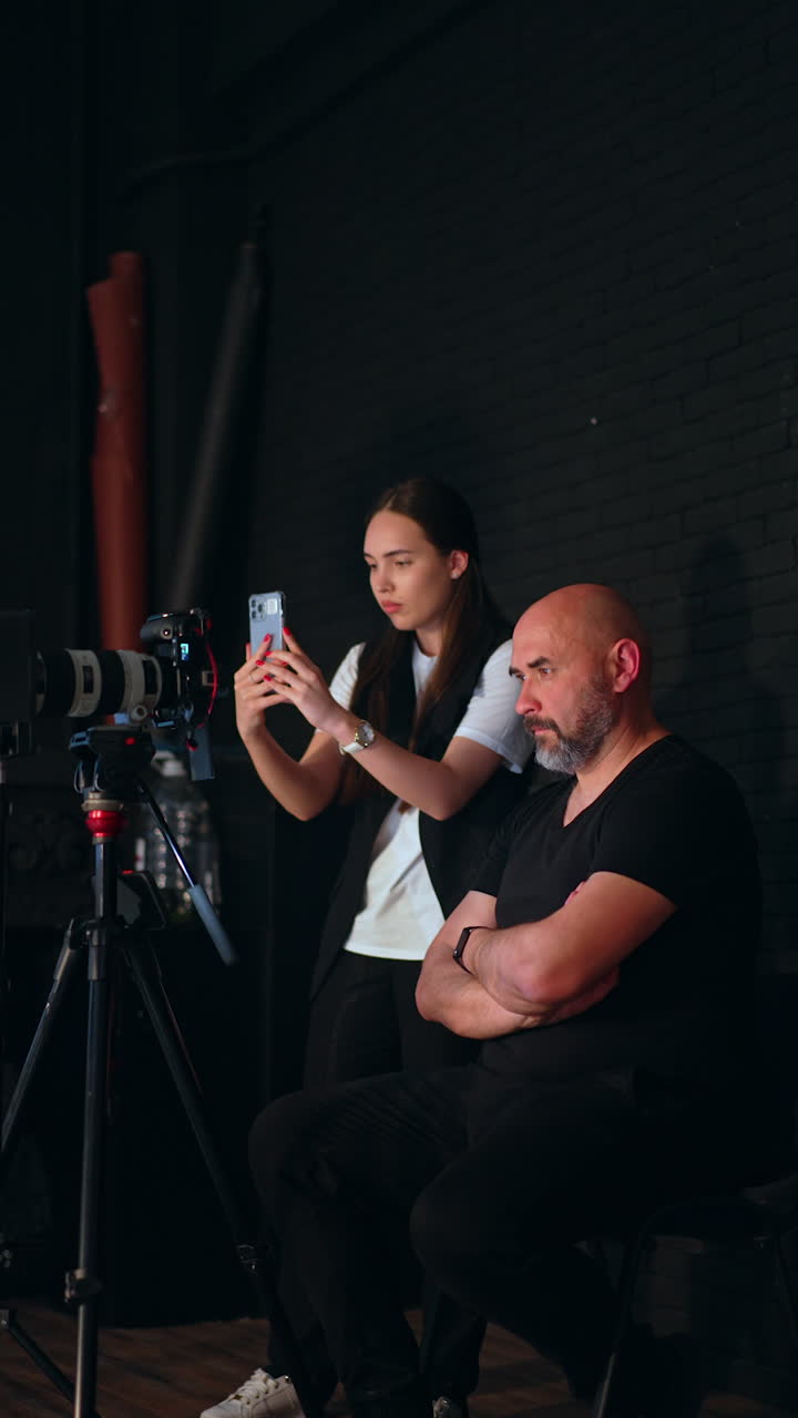 Cameraman sits with his arms crossed on chest looking ahead. Woman standing beside uses her phone for taking photos or video. Studio footage. Vertical video.