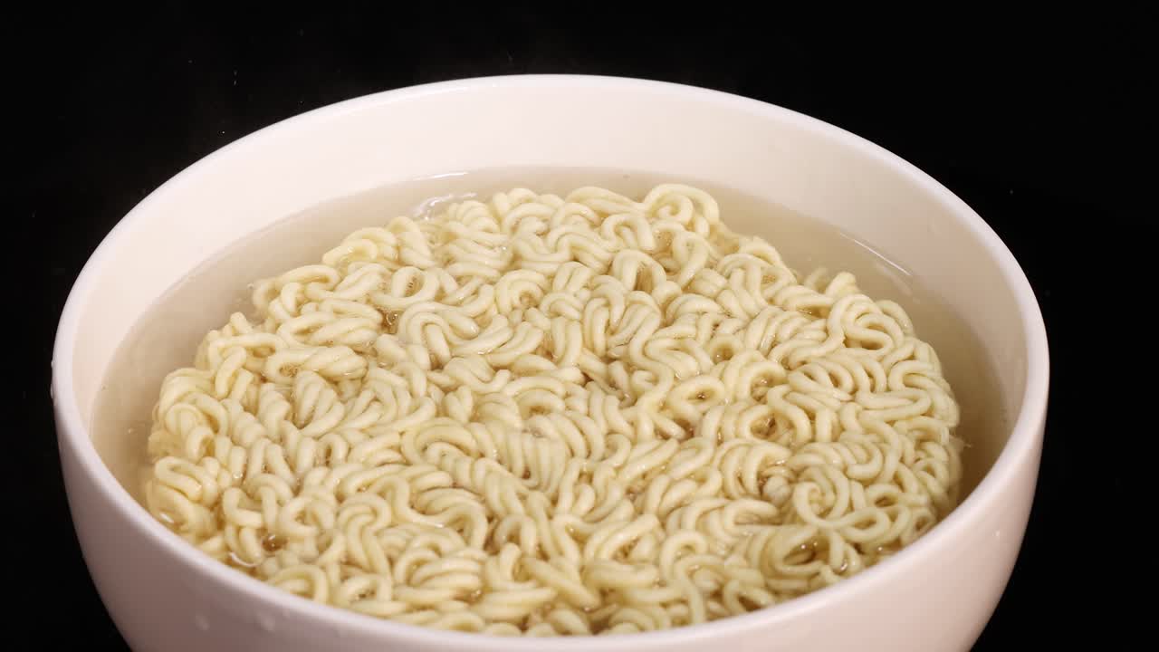 Overhead view of instant noodles in a bowl absorbing hot water, expanding and softening