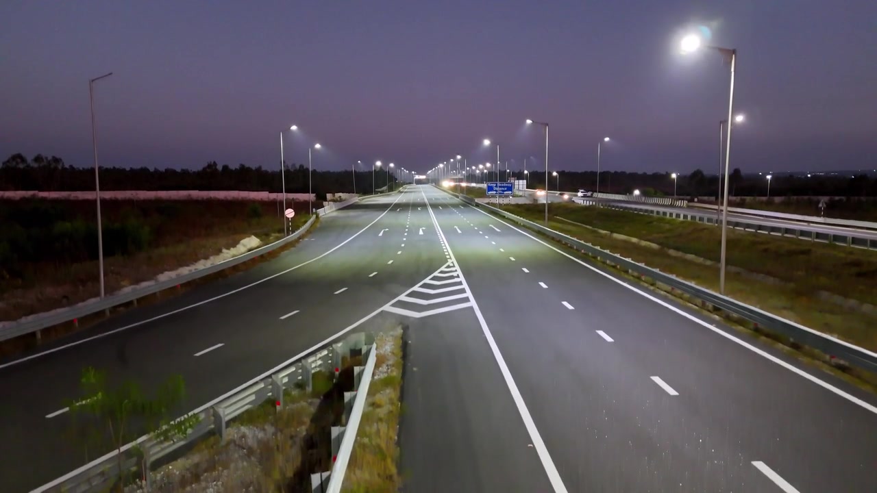Ripresa con drone che cattura la superstrada Bengaluru-Chennai al tramonto, con corsie autostradali illuminate, veicoli in movimento e lunghi schemi stradali simmetrici che attraversano il paesaggio notturno.