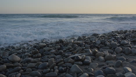 Toma-Panorámica-De-Las-Olas-Rompiendo-En-La-Costa-Rocosa-De-La-Playa-De-Mondo-Con-La-Puesta-De-Sol-Detrás-De-Una-Montaña-En-El-Fondo