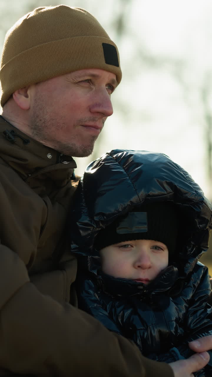 Father and son sitting on a bench, with the father resting his head gently on his son's head, with the father holding his son close, with trees in the background