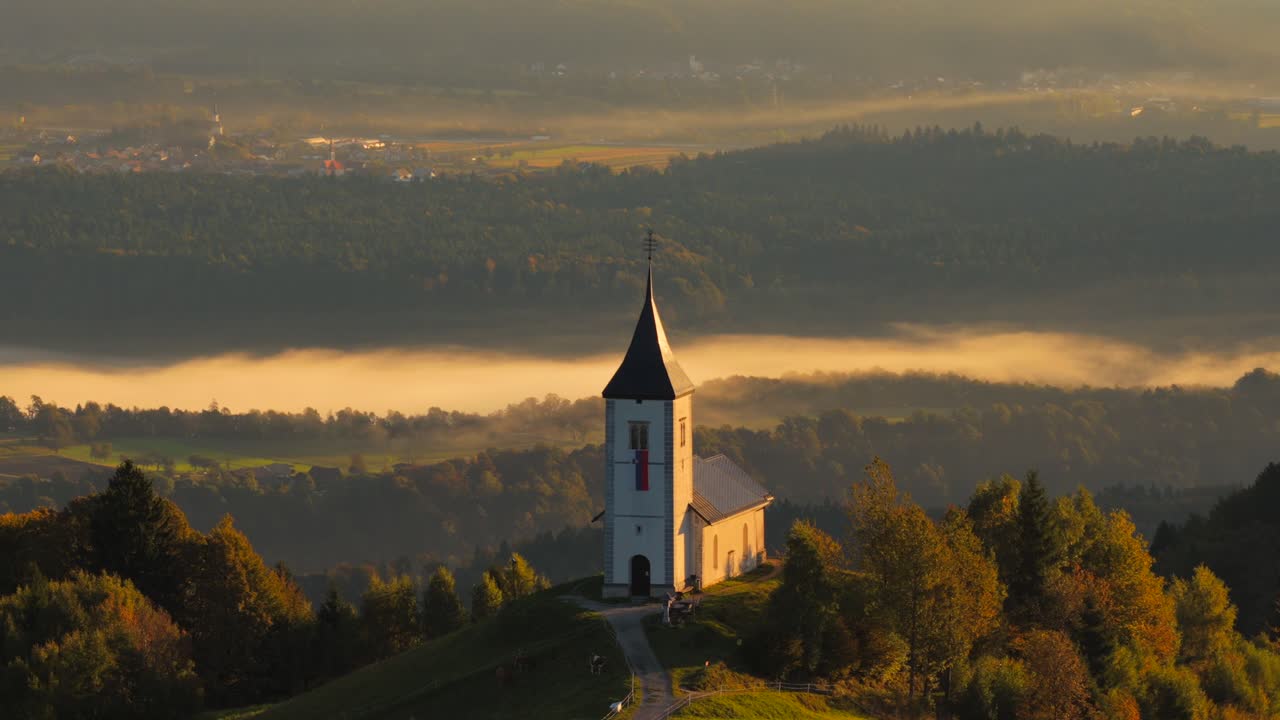 Aerial establishing view of the Church of St. Primus and Felician at Jamnik, Slovenia, surrounded by autumn hills and backed by the Julian Alps, showcasing peaceful alpine scenery at sunrise