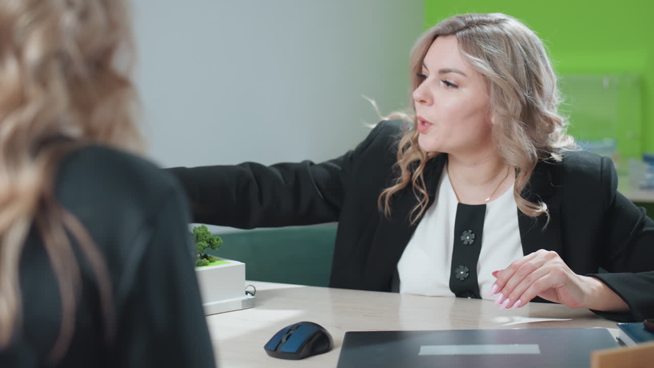 Business expert gesturing with hand while explaining details of building model to client during focused office conversation, dressed professionally, sitting across desk with bright background