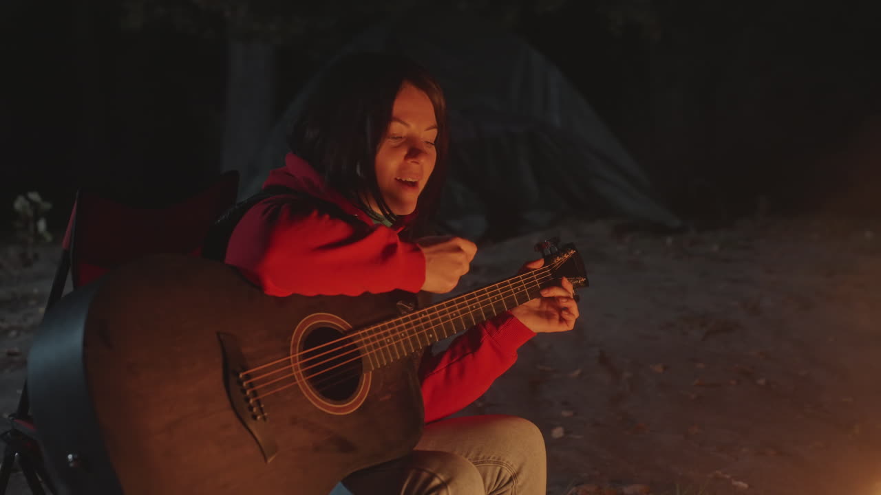 Notte in campeggio, una donna suona la chitarra e canta accanto a un falò, inquadratura ravvicinata su mani e viso, scintille che salgono dalla fiamma, sagoma della tenda sullo sfondo, delicato strimpellare ed espressione vocale, bagliore della lanterna.