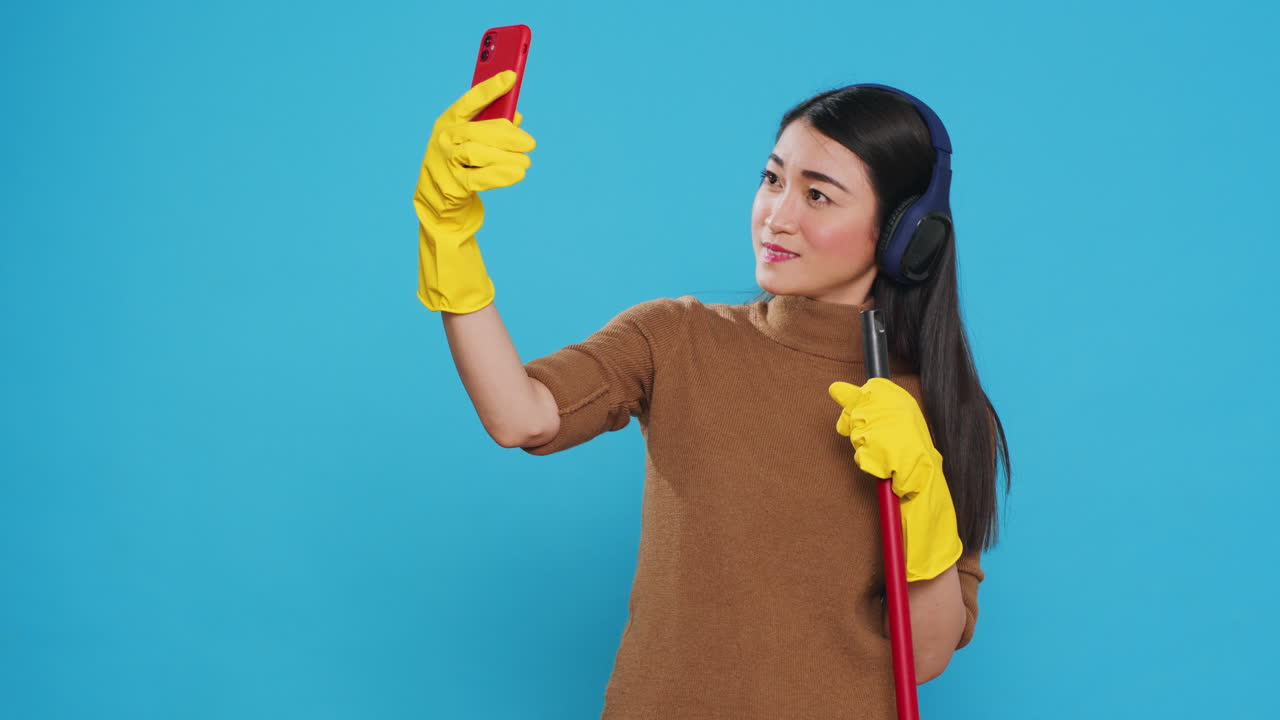 Cheerful housekeeper holding cleaning broom while taking selfie with smartphone