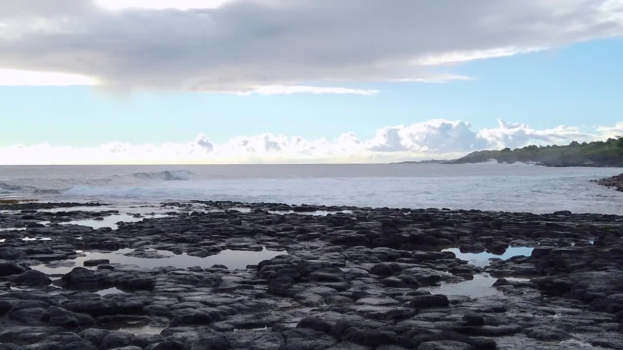 hd 슬로우 모션 하와이 카우아이 정적 파도가 언덕이 있는 용암 바위를 향해 왼쪽에서 오른쪽으로 굴러 들어오고 부분적으로 흐린 하늘이 있는 프레임 오른쪽의 거리에서 분출하는 혼 블로우홀