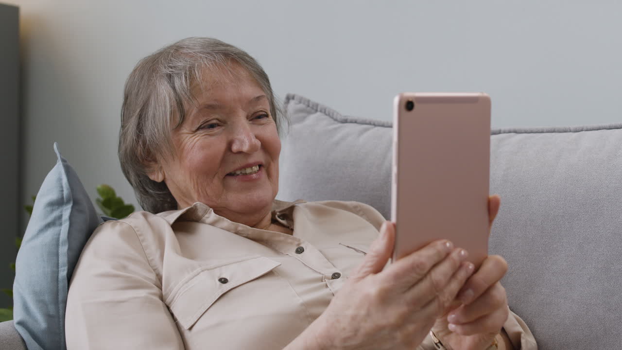 mujer mayor feliz que tiene una videollamada a través de una tableta mientras está acostada en el sofá en casa