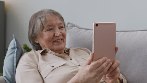 mujer mayor feliz que tiene una videollamada a través de una tableta mientras está acostada en el sofá en casa