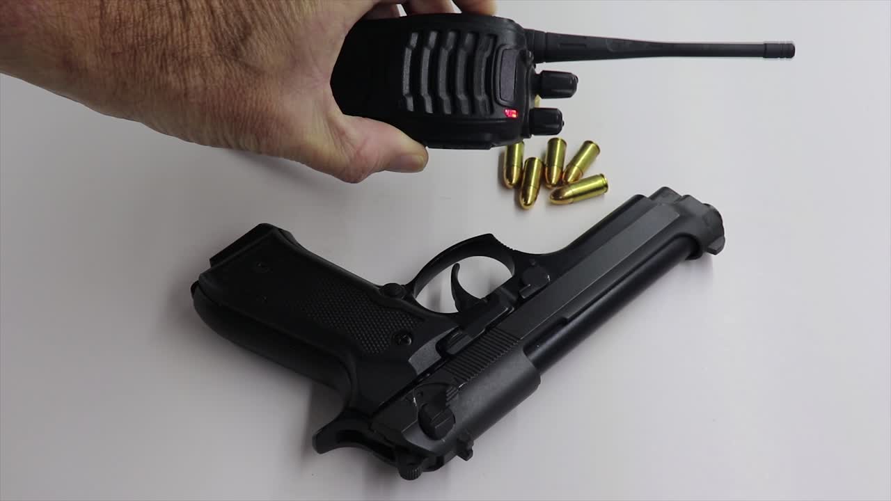 persona tecleando el micrófono en una radio de mano negra luego recoge un arma de fuego negra dejando algunas balas detrás