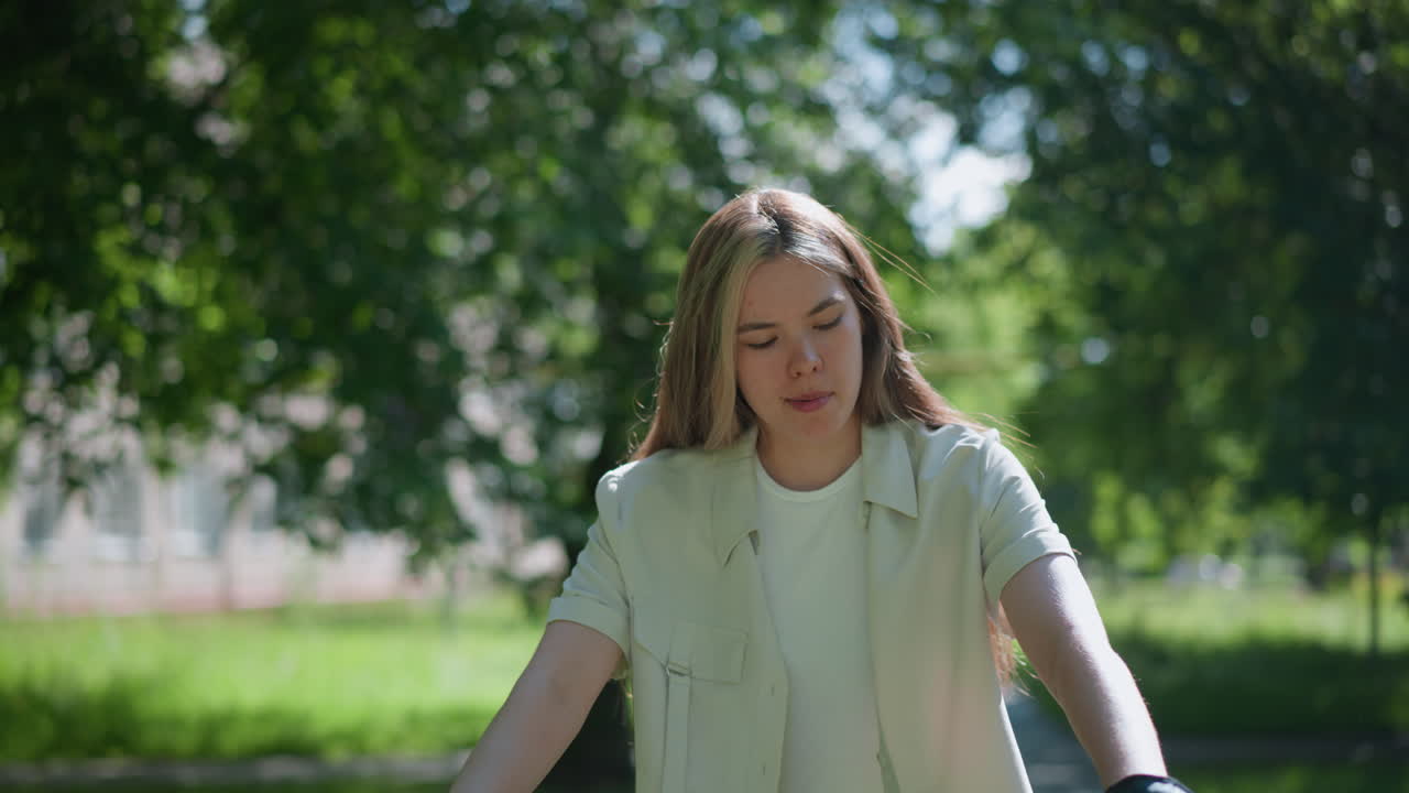 초점을 맞춘 젊은 여성은 자전거에 앉아 자전거 장갑을 입고 손을 집중적으로 쳐다보고 있습니다. 배경은 울창하고 바람에 부힌 잎과 진정된 자연 환경을 보여줍니다.