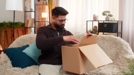 indian man on sofa opens box and shakes head after discovering wrong item in online delivery order