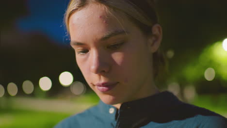 primer plano de una mujer joven en un tranquilo parque al aire libre por la noche, mostrando una expresión pensativa bajo el suave resplandor de las luces, el fondo incluye vegetación borrosa y iluminación suave
