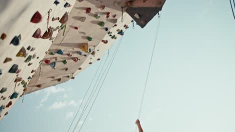 Bottom-view:-an-athletic-girl-climbs-to-the-required-height-on-a-climbing-wall,-and-then-climbs-down-using-a-safety-net.-A-blonde-girl-goes-down-using-ropes-during-a-rock-climbing-lesson