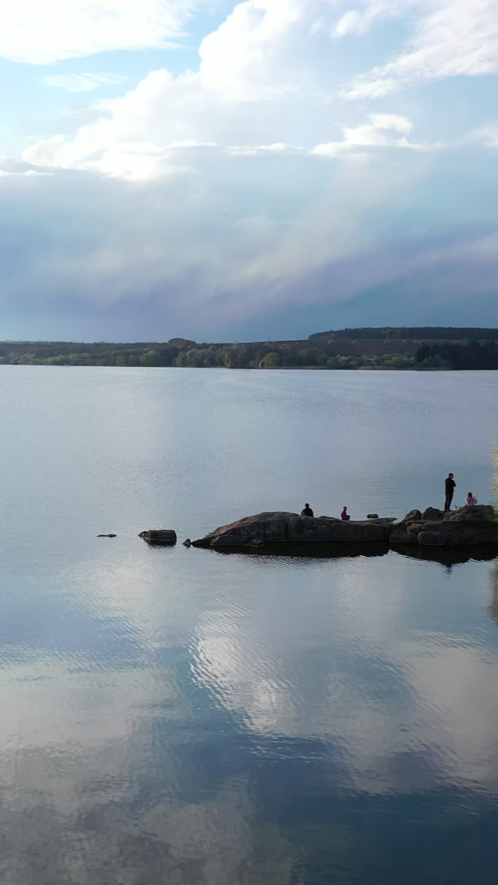 Beautiful river on nature landscape in spring. People rest near the water on stones in springtime. Blooming trees on a bank of the river. Vertical video