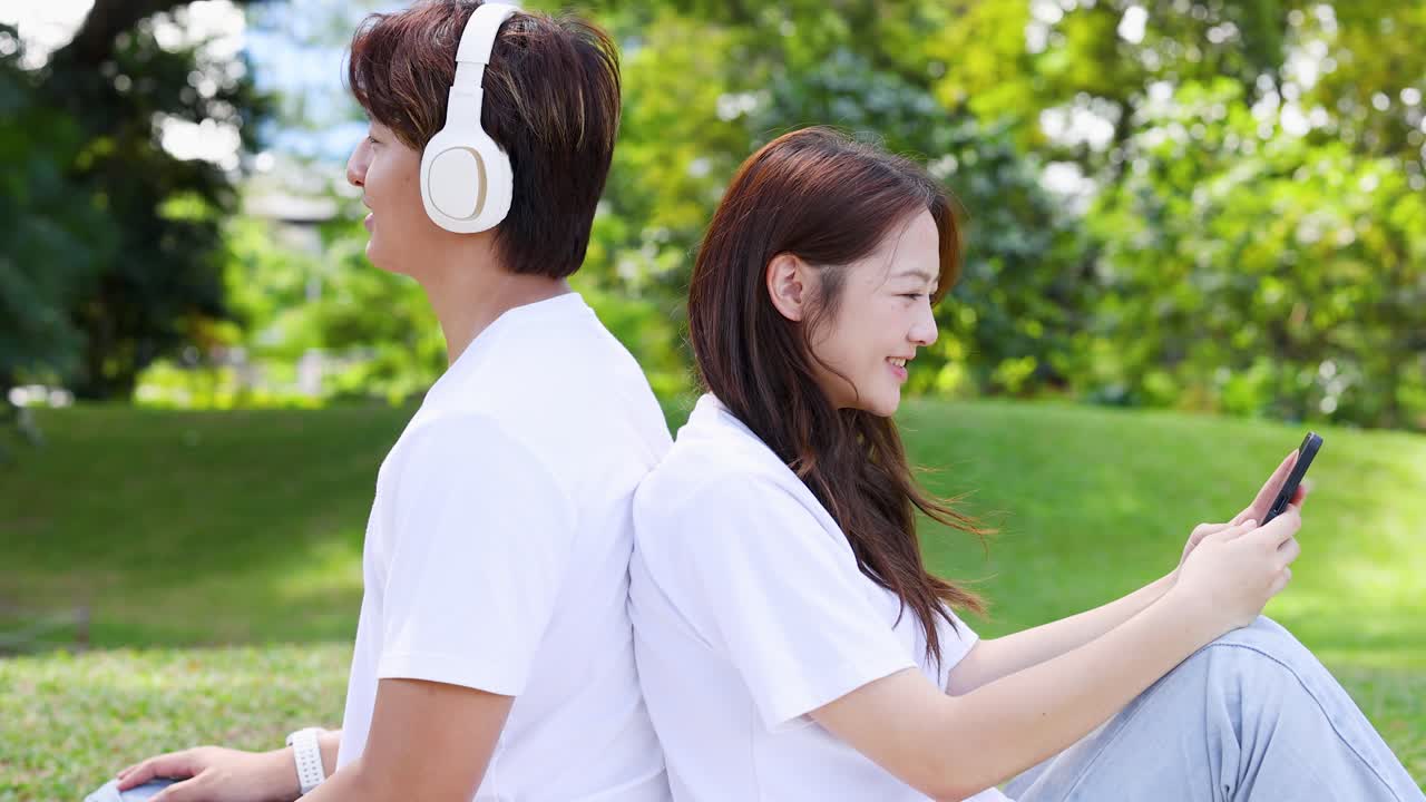 若いアジア人のカップルが、芝生の上で背中合わせに座り、ヘッドホンを着けてスマートフォンを使っています。