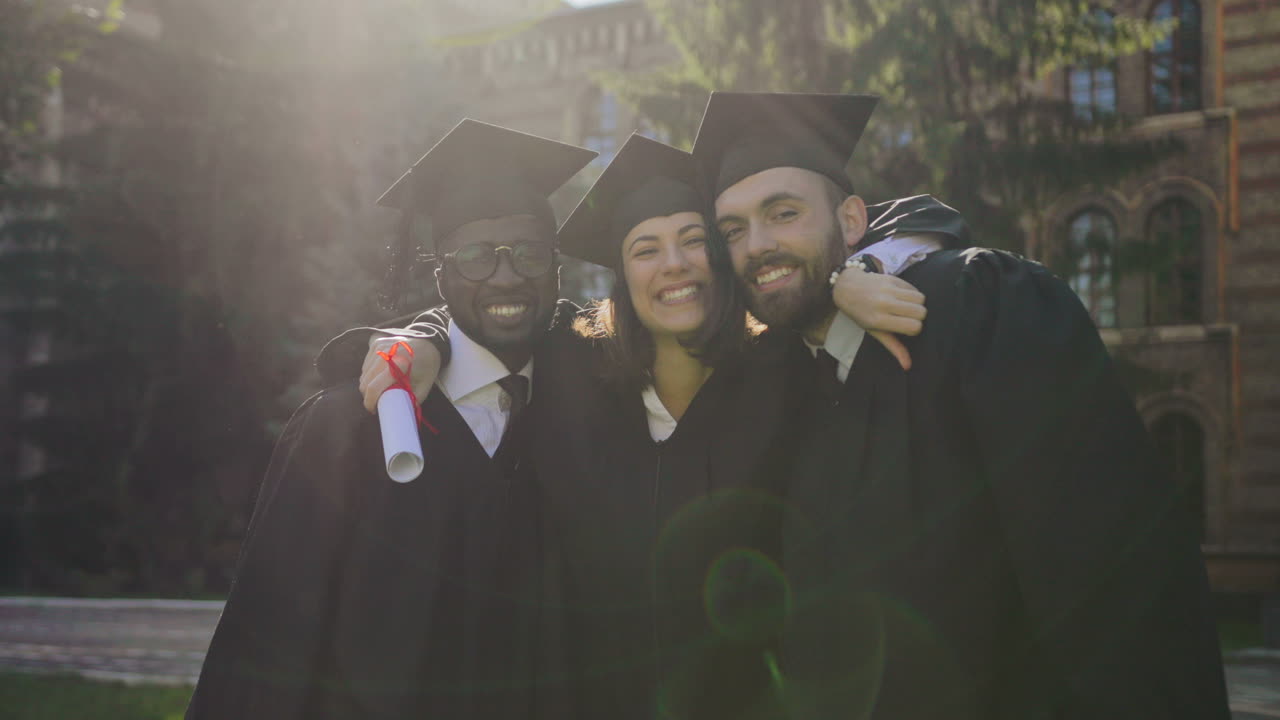 Portrait shot of young woman and men in traditional clothes and caps posing to the camera and hugging on their graduation day