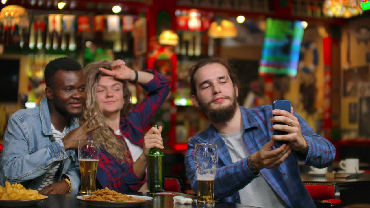 Un gruppo di amici, un uomo e due ragazze, si scattano un selfie in un bar ridendo e sorridendo. Trascorrono del tempo con gli amici in un bar.