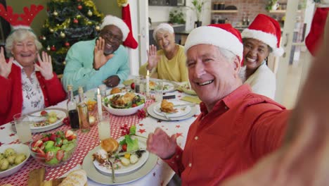 felices amigos mayores diversos en sombreros de santa tomando selfies y saludando en la mesa de la cena de navidad