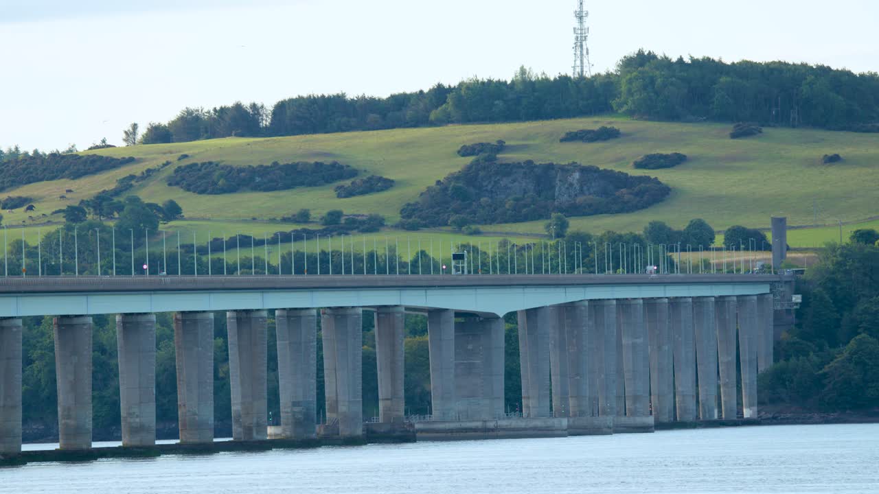 Truck crosses large river bridge in rural landscape with rolling hills under soft daylight