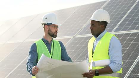 two caucasian and african american farm solar engineers talking while looking at blueprint on solar plantation