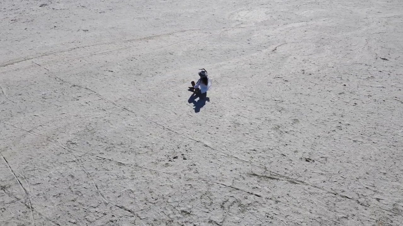 Lone individual wearing hat sits on cracked dry earth under bright sunlight conveying solitude and