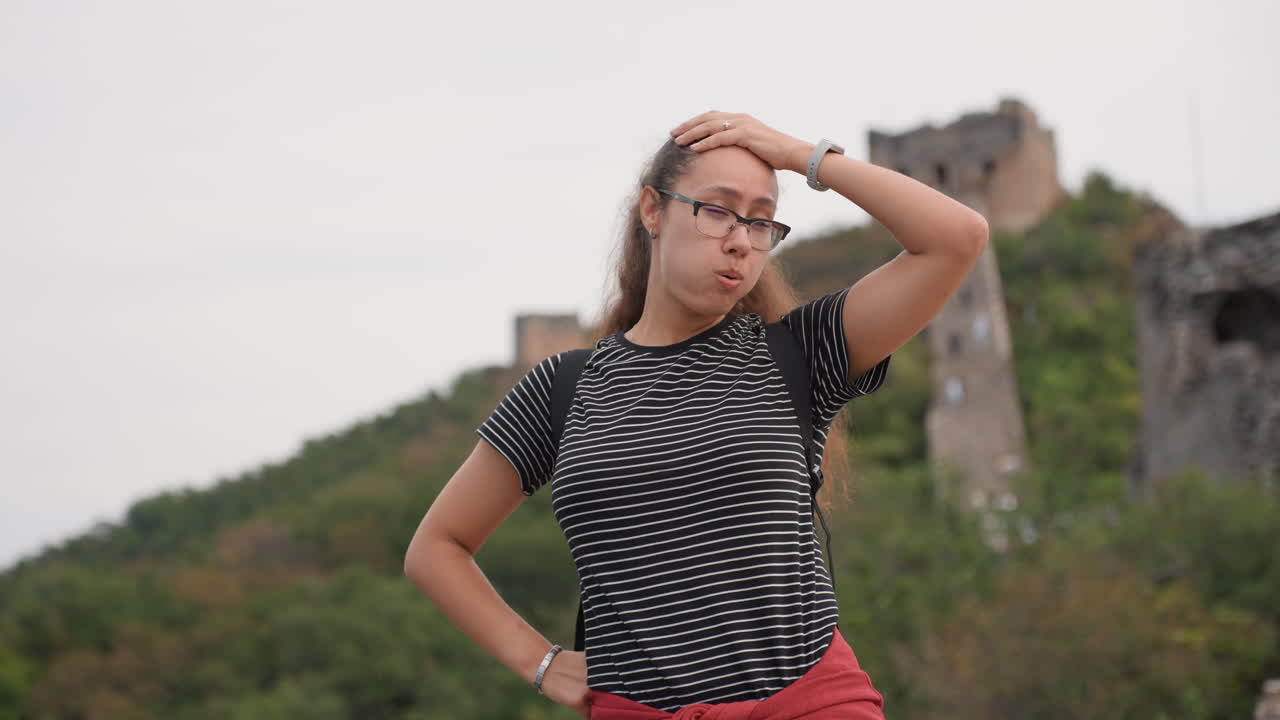 Confident young woman posing at ruins, hands on hips and glasses reflecting overcast sky striped shirt and red pants contrast stone castle towers behind, breeze tossing ponytail, solo traveler vibe